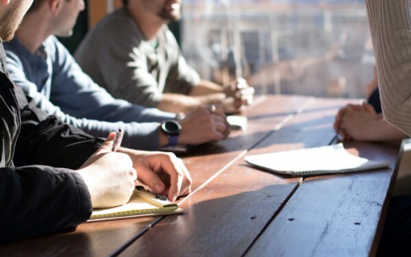 three people sitting at table with pens in their hand and one person standing up on opposite side of table with paper in front of them on table
