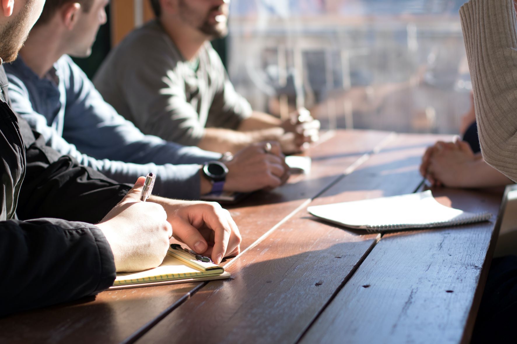 three people sitting at table with pens in their hand and one person standing up on opposite side of table with paper in front of them on table
