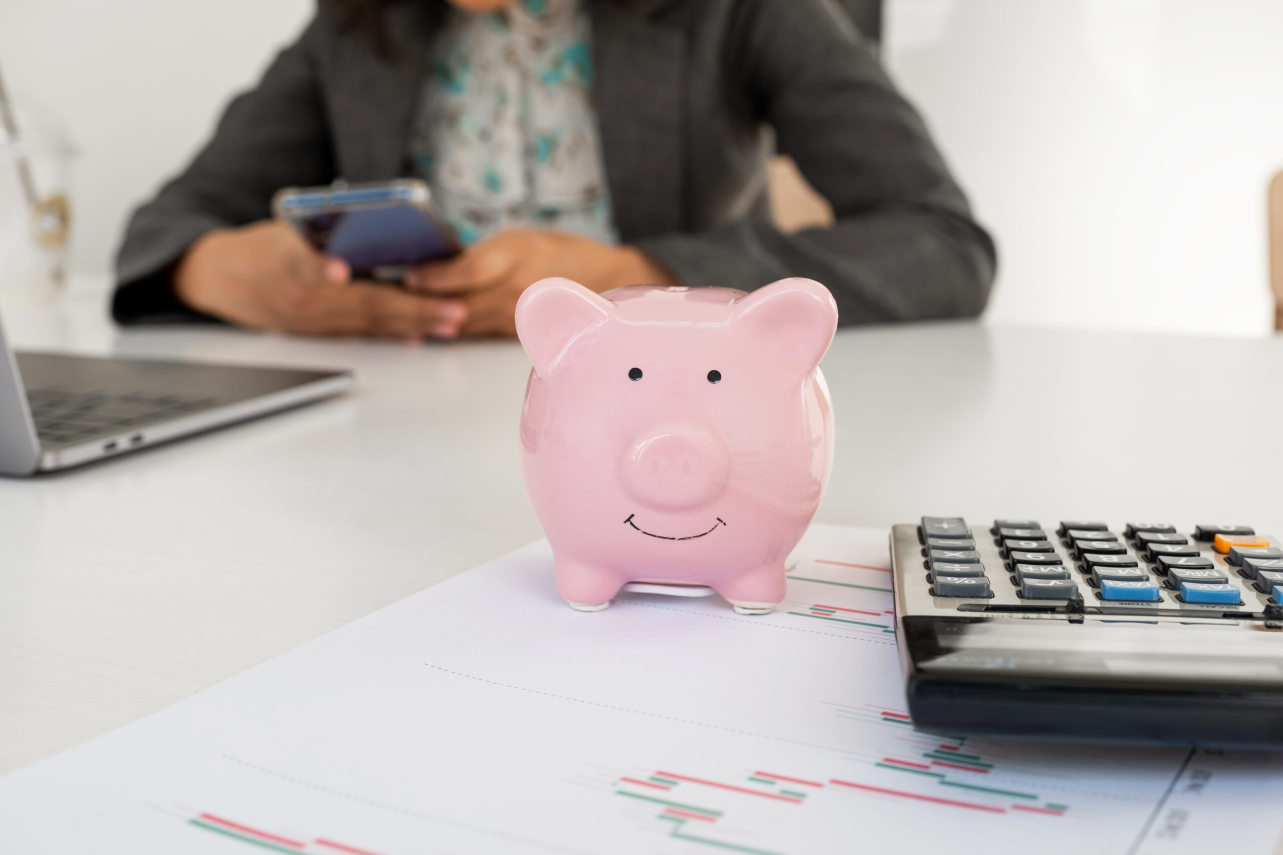 person holding cell phone in their hands at desk with pink piggy bank calculator and papers