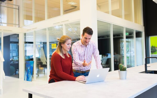 woman sitting at white desk with man standing on her right pointing at computer screen