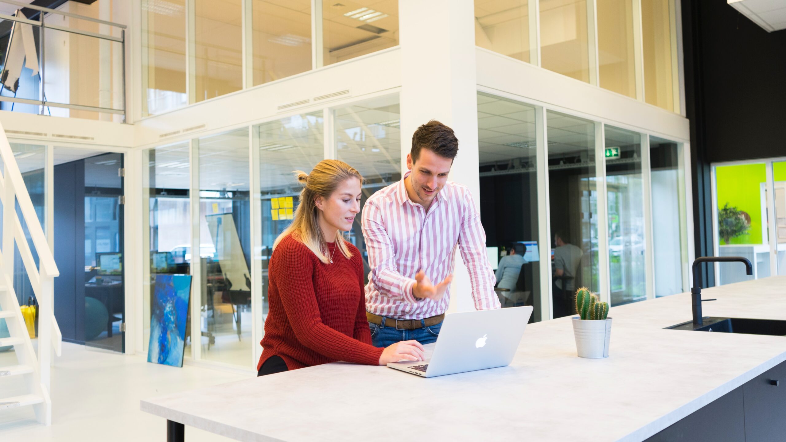 woman sitting at white desk with man standing on her right pointing at computer screen