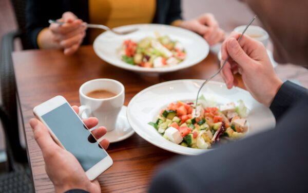 man in suit jacket holds his cell phone while holding a fork above his salad sitting across from a colleague eating the same salad
