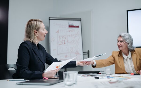 younger blonde woman handing older woman paper while sitting at white conference table