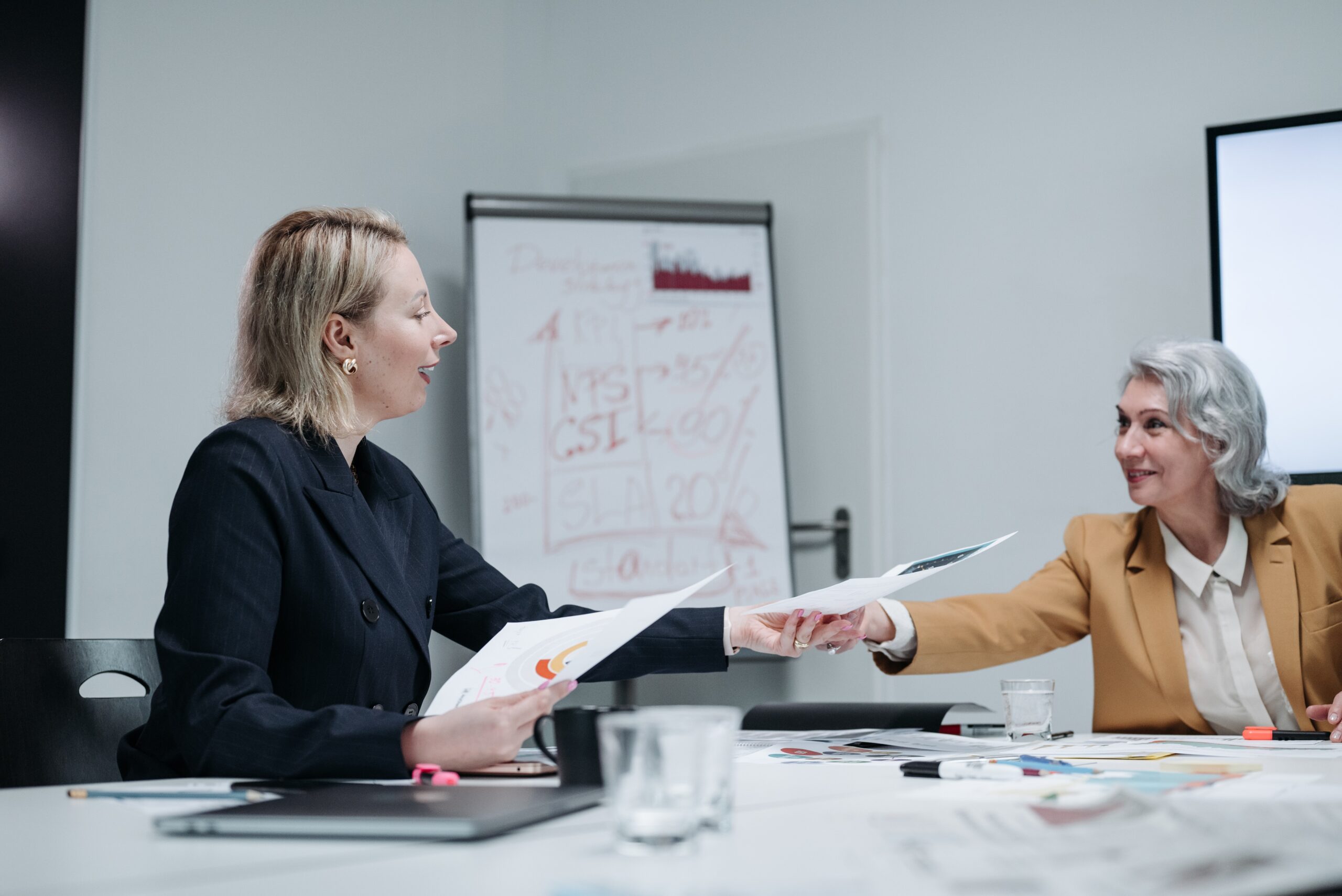younger blonde woman handing older woman paper while sitting at white conference table