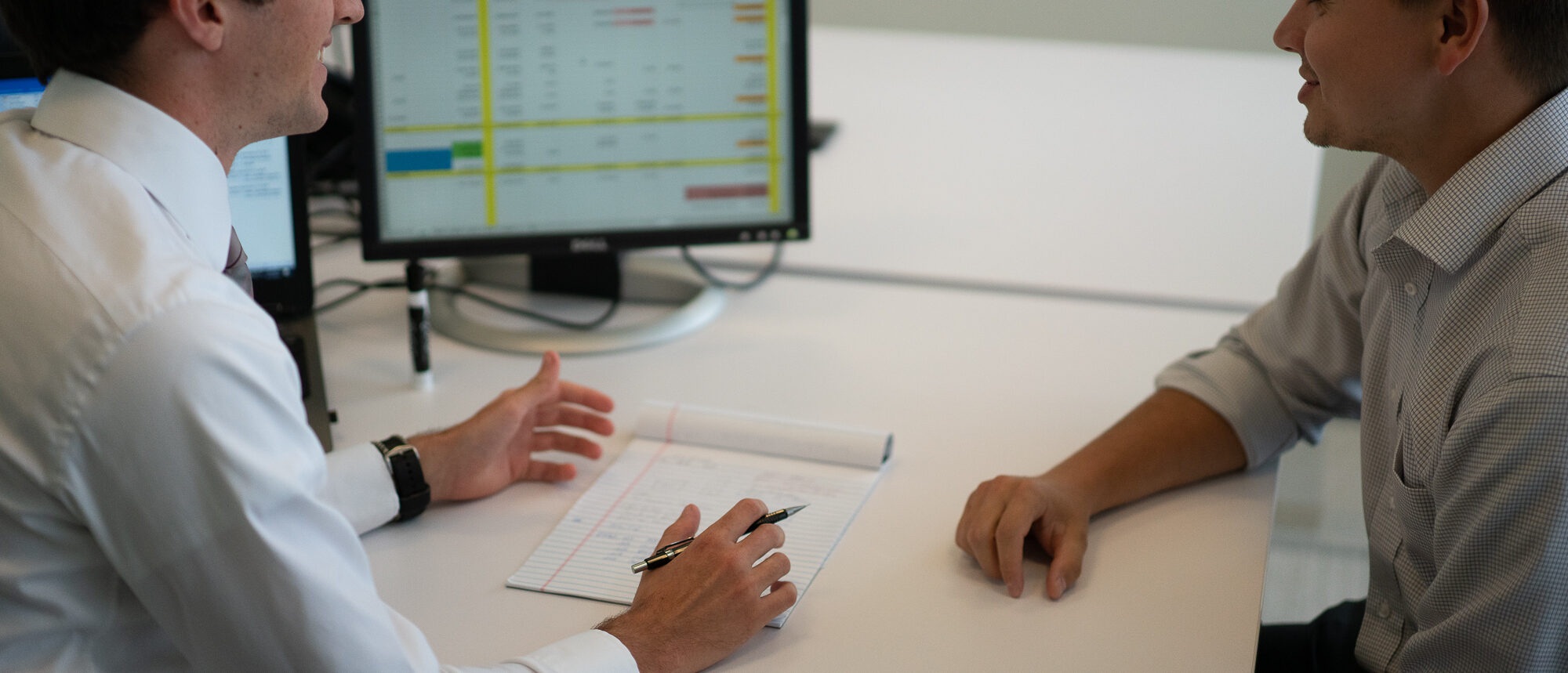 Two men sit at desk discussing a spreadsheet on a computer screen